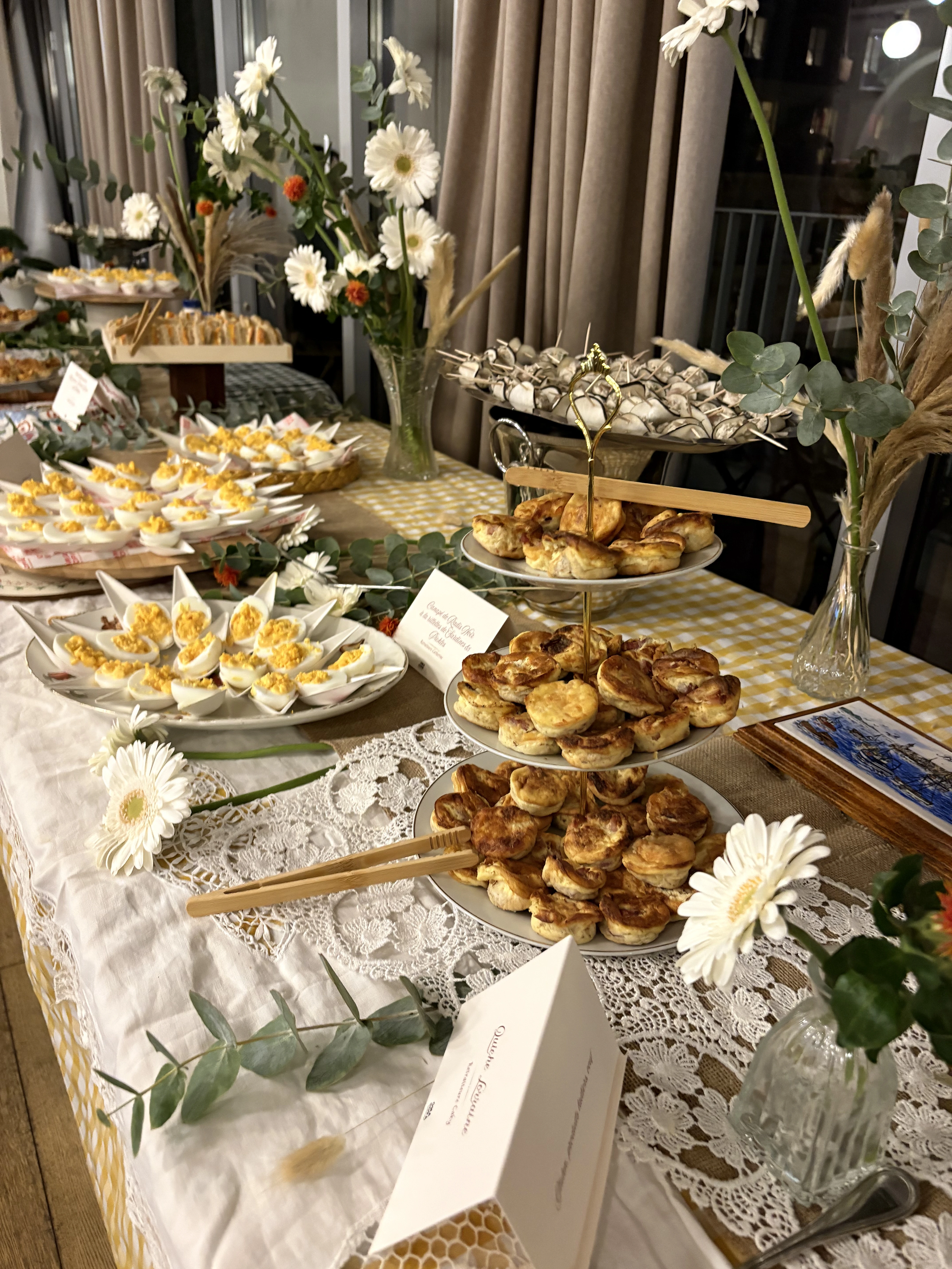 Table de buffet avec des assiettes à plusieurs niveaux garnies de pâtisseries, d'œufs mimosa, de marguerites blanches dans des vases et de verdure décorative sur des nappes en dentelle et à carreaux jaunes.