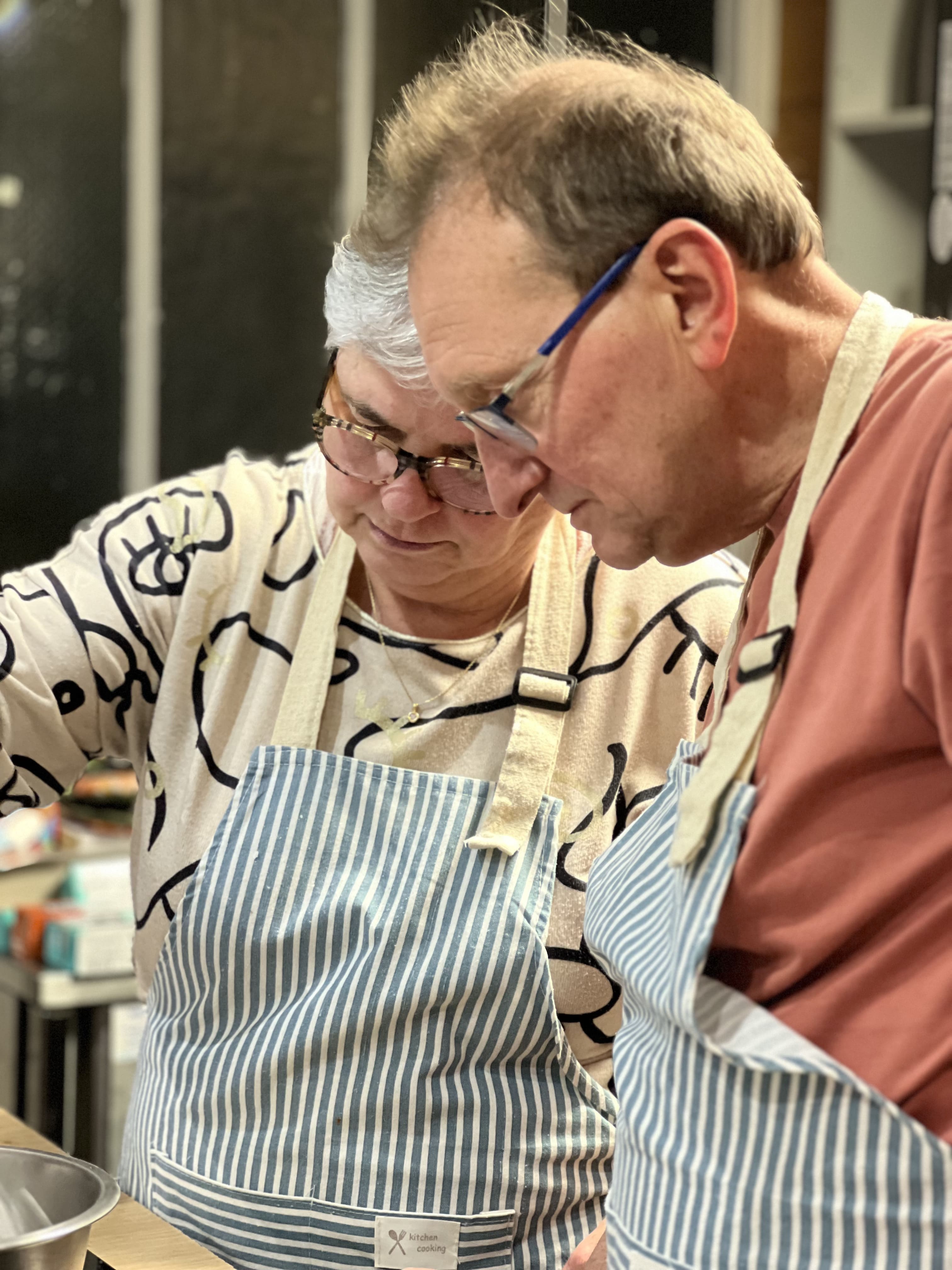 Two older adults wearing striped aprons, concentrating on cooking together in a kitchen.