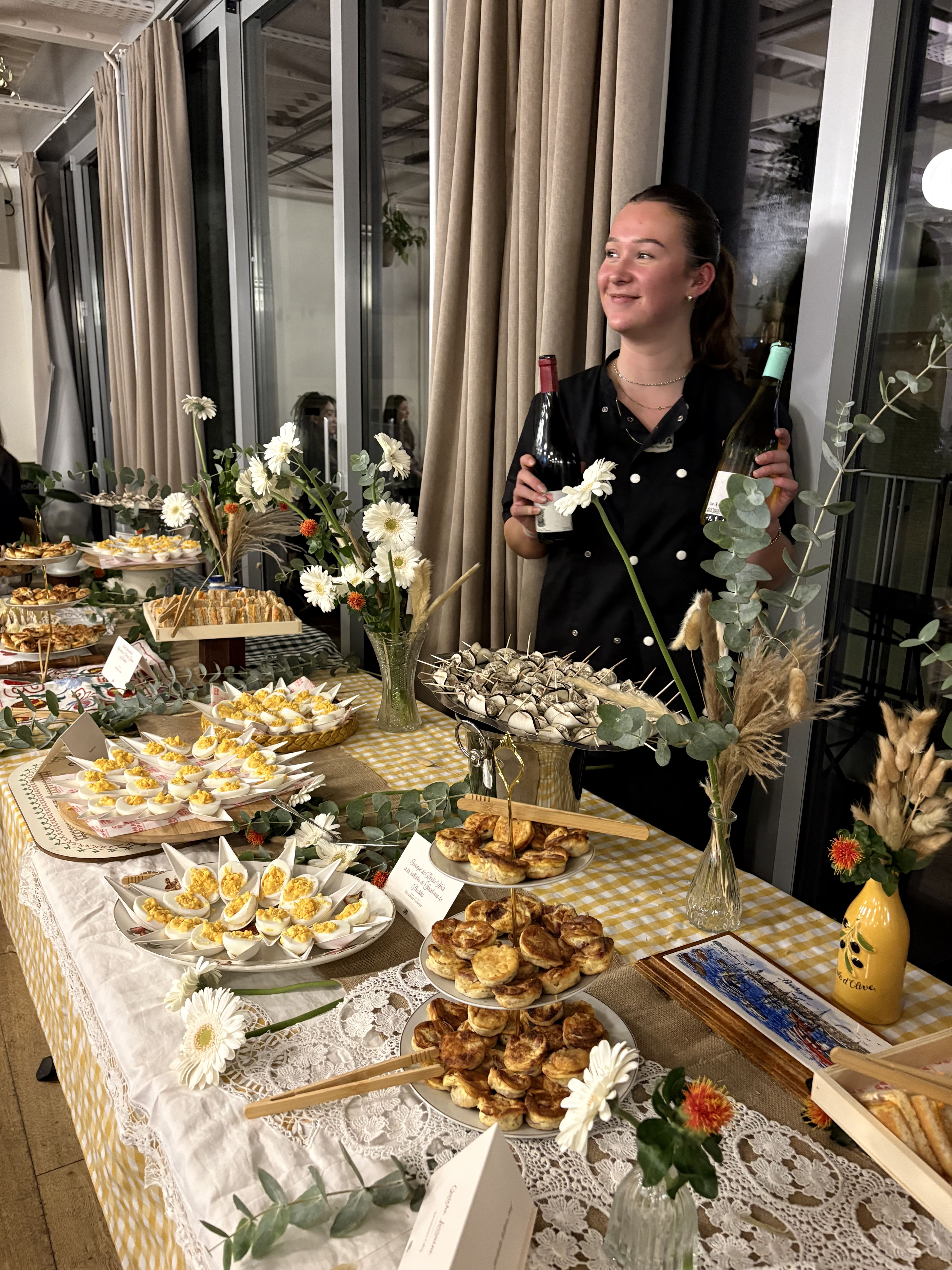 Woman in black holding two wine bottles behind a decorated table with various appetizers and flowers.