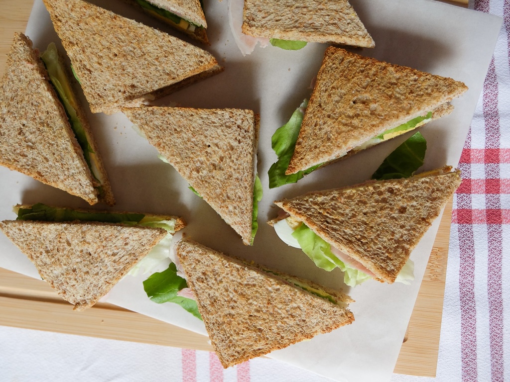 Triangle sandwiches with whole wheat bread filled with lettuce and deli meat arranged on parchment paper over a wooden board.