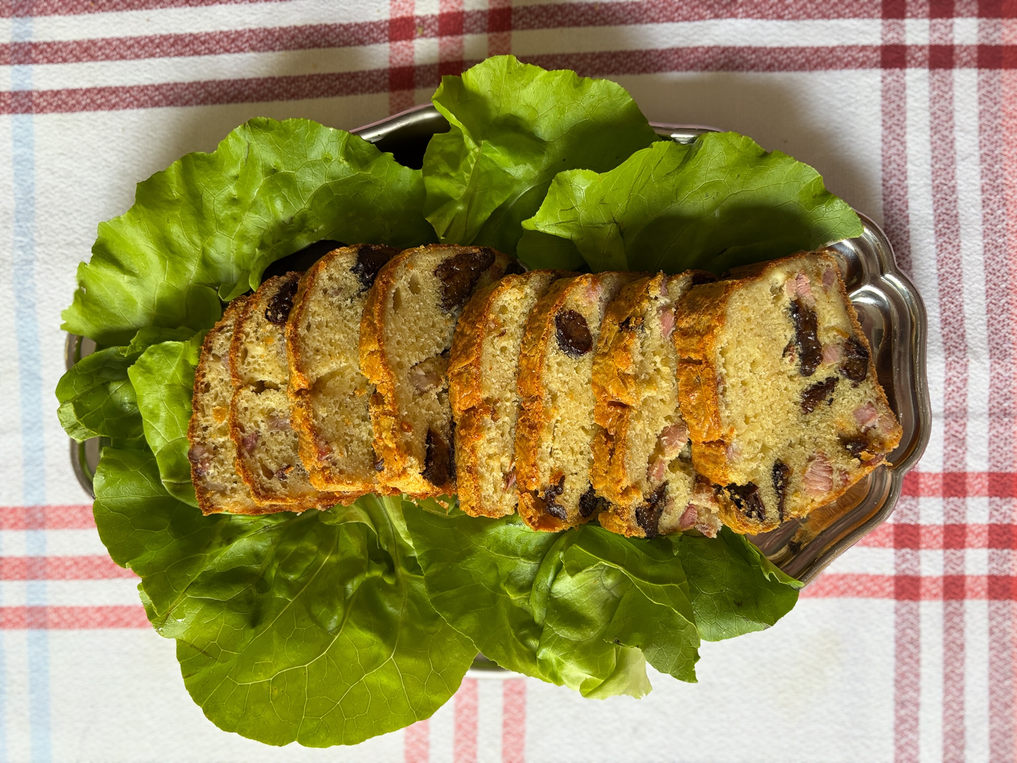 Sliced fruit and nut loaf arranged on green lettuce leaves on a silver platter over a red and blue checkered tablecloth.