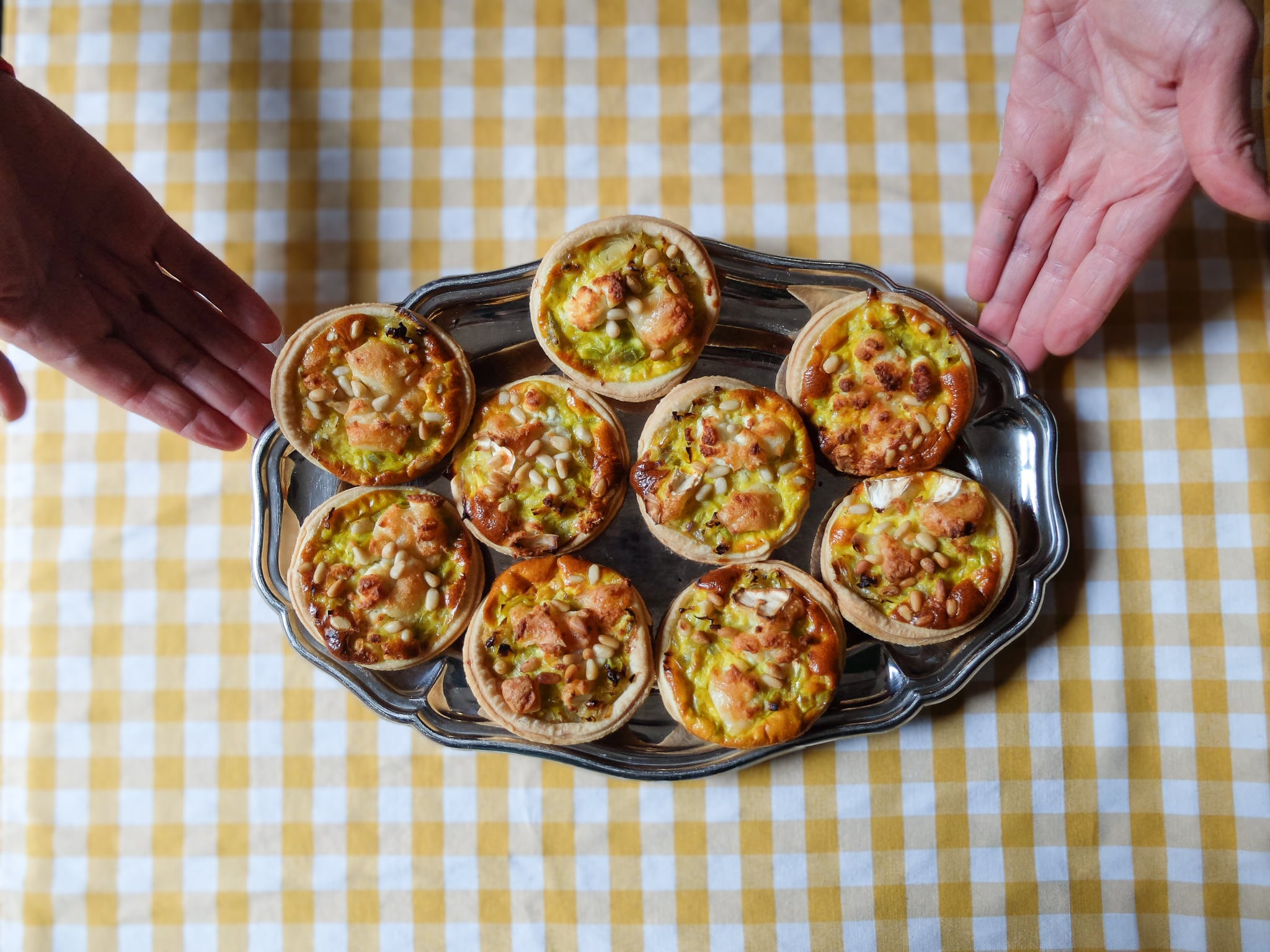 Silver tray holding eight small savory tarts topped with pine nuts on a yellow and white checkered tablecloth, with two hands on either side.