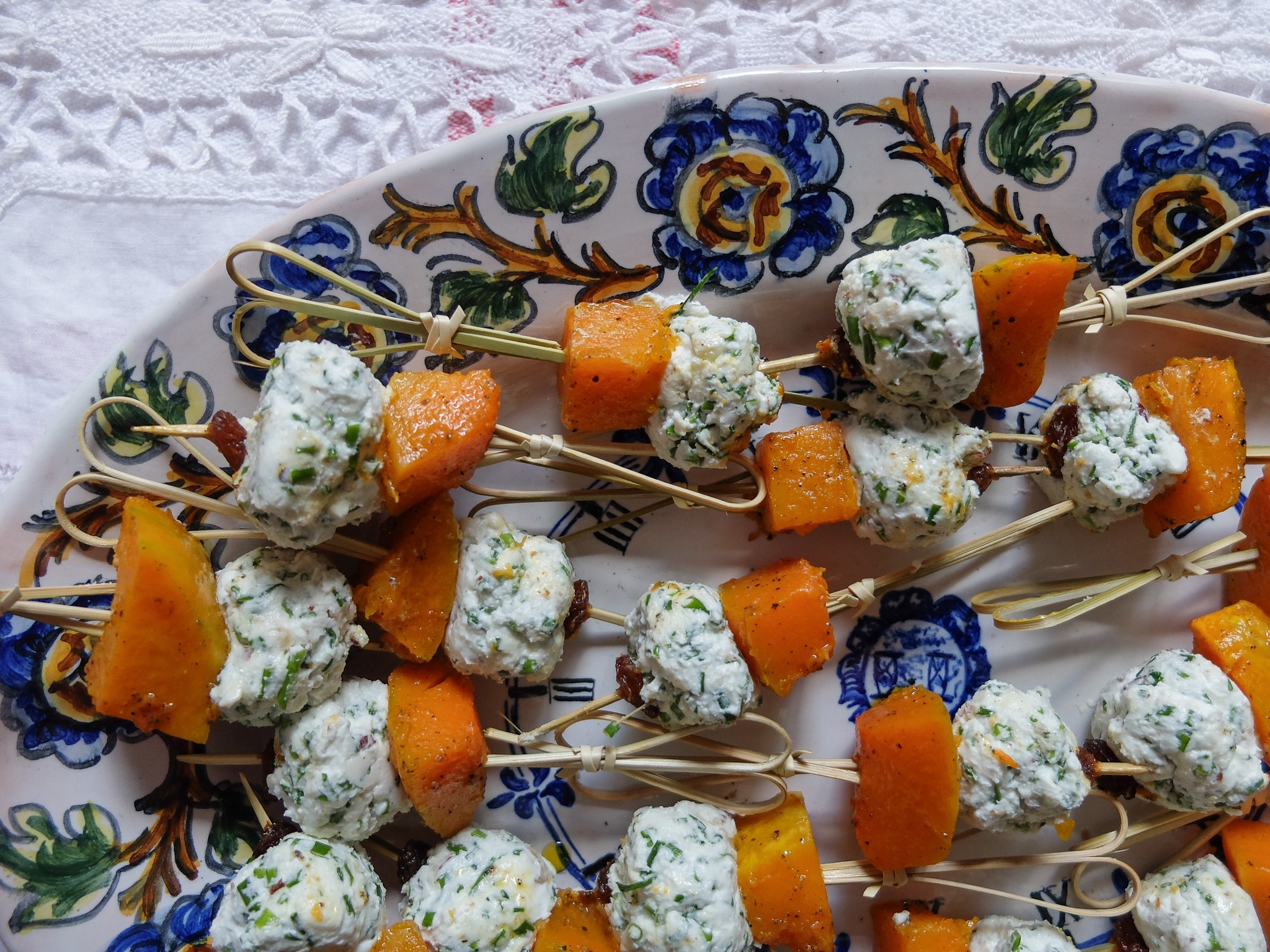 Skewers with chunks of orange roasted pumpkin and white herb-infused cheese balls on a decorative plate with blue and green floral patterns.
