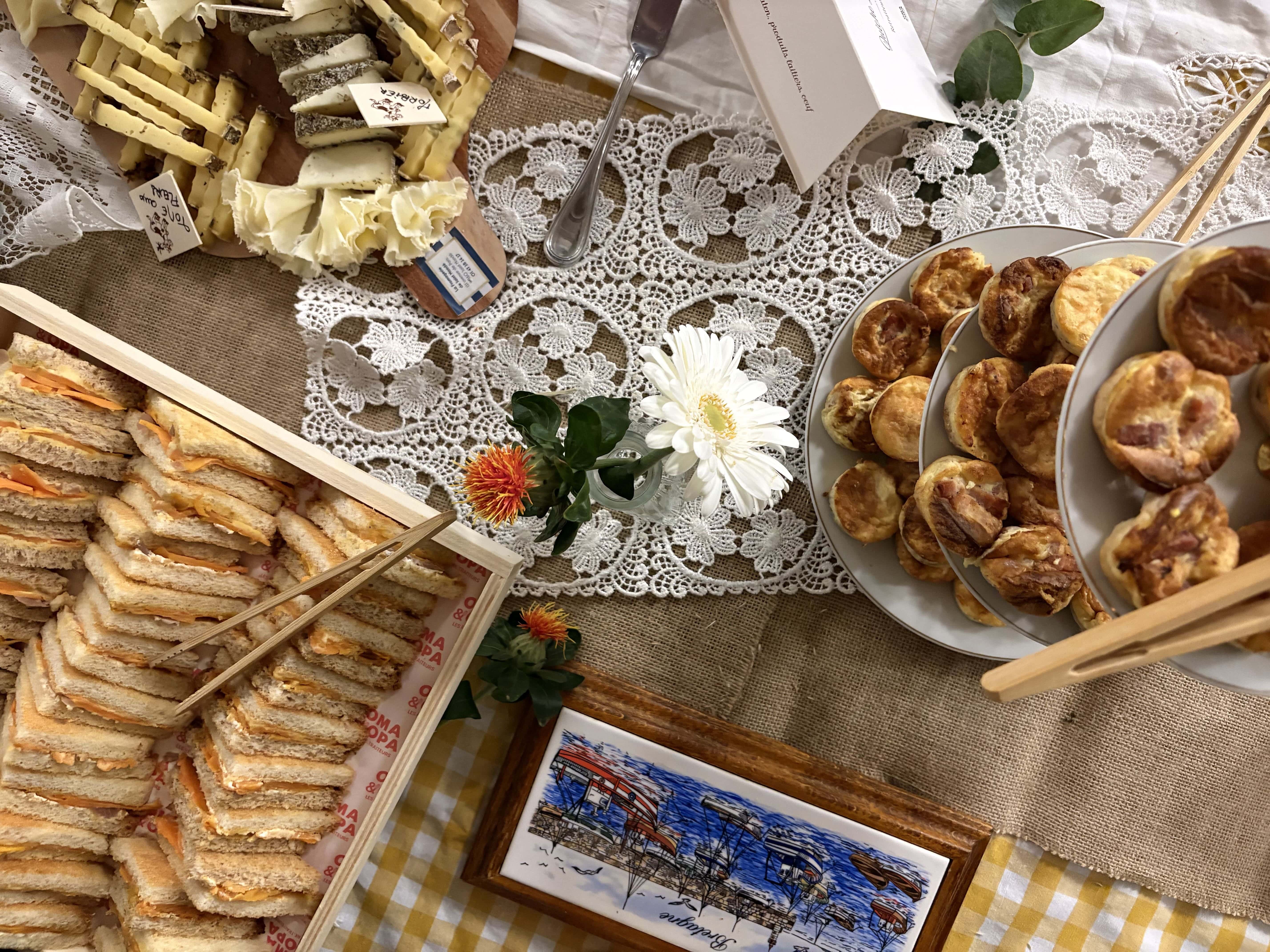 Table with assorted finger foods including stacked tea sandwiches, small savory pastries on tiered plates, cheese platter with labels, lace tablecloth, and small vases with white and orange flowers.