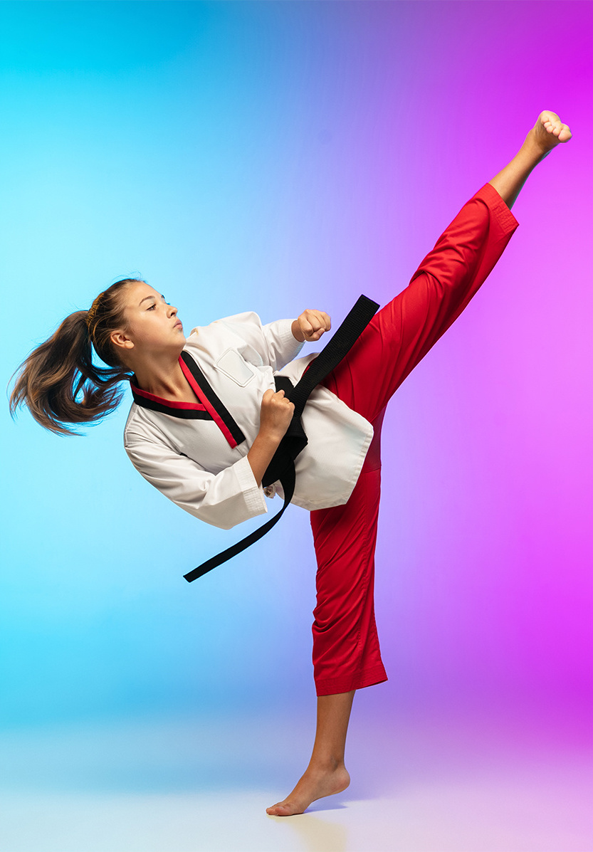 A girl in a red and white karate uniform doing a vertical kick against a blue and pink background