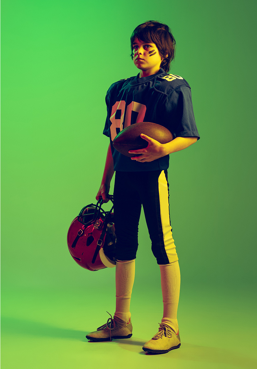 A boy in a football uniform holding a football and a helmet against a green background