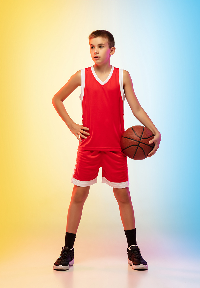 A boy in a red basketball uniform holding a basket ball with his hand on his hip against a yellow and blue background