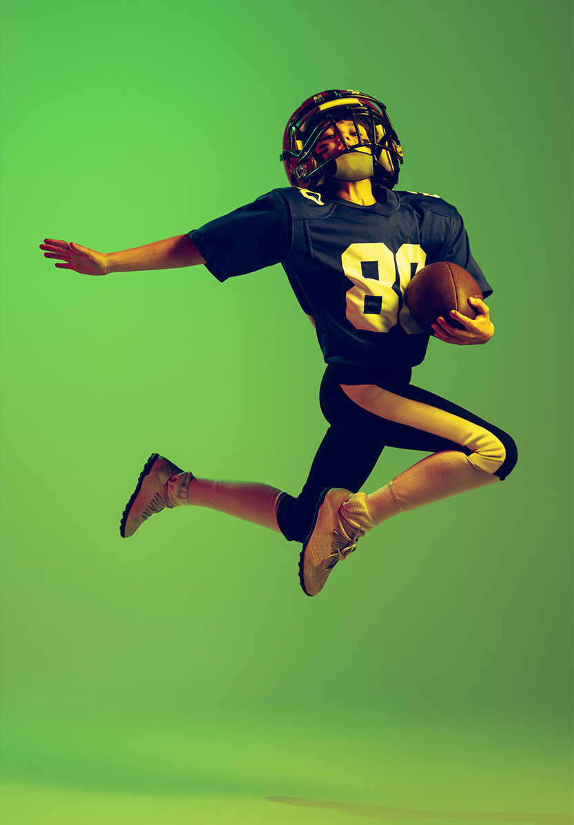 A boy in a football uniform jumping through the air with a football against a green background