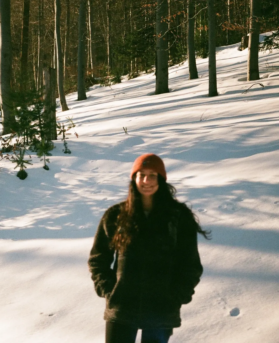 Person wearing a red beanie and dark coat standing in a snowy forest with tall trees casting long shadows.