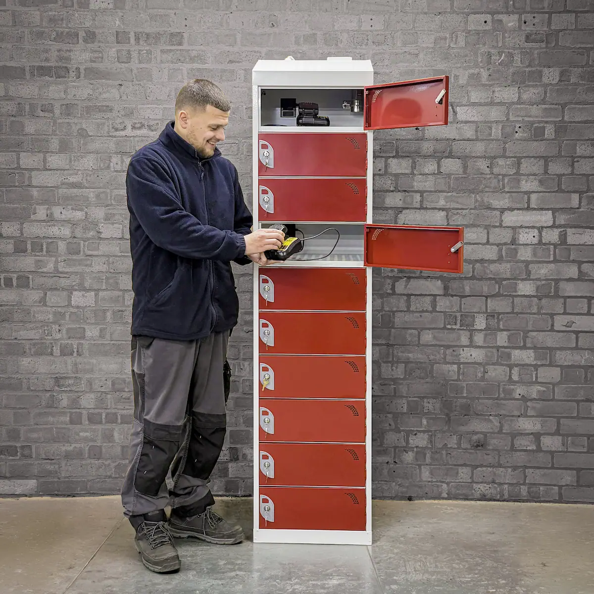 Man handling a charging device near an open red locker in a tall white locker unit against a brick wall.