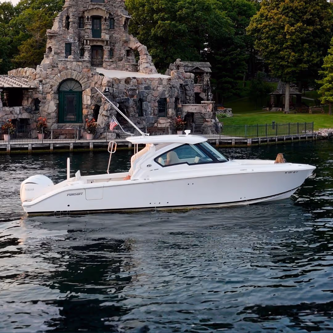 White Pursuit motorboat floating on water near a stone building and green trees.