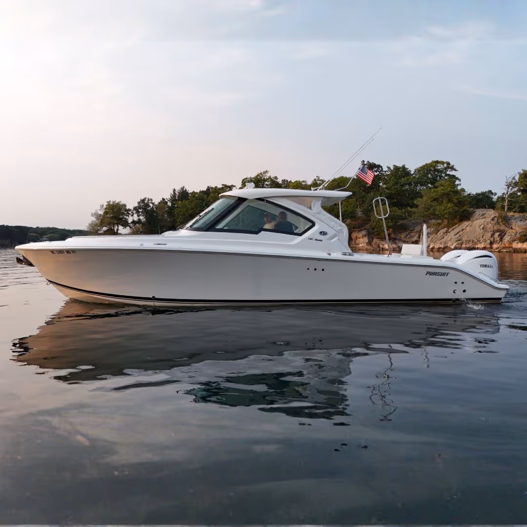 White Pursuit powerboat with Yamaha engines sailing on calm water near a rocky shore with trees.