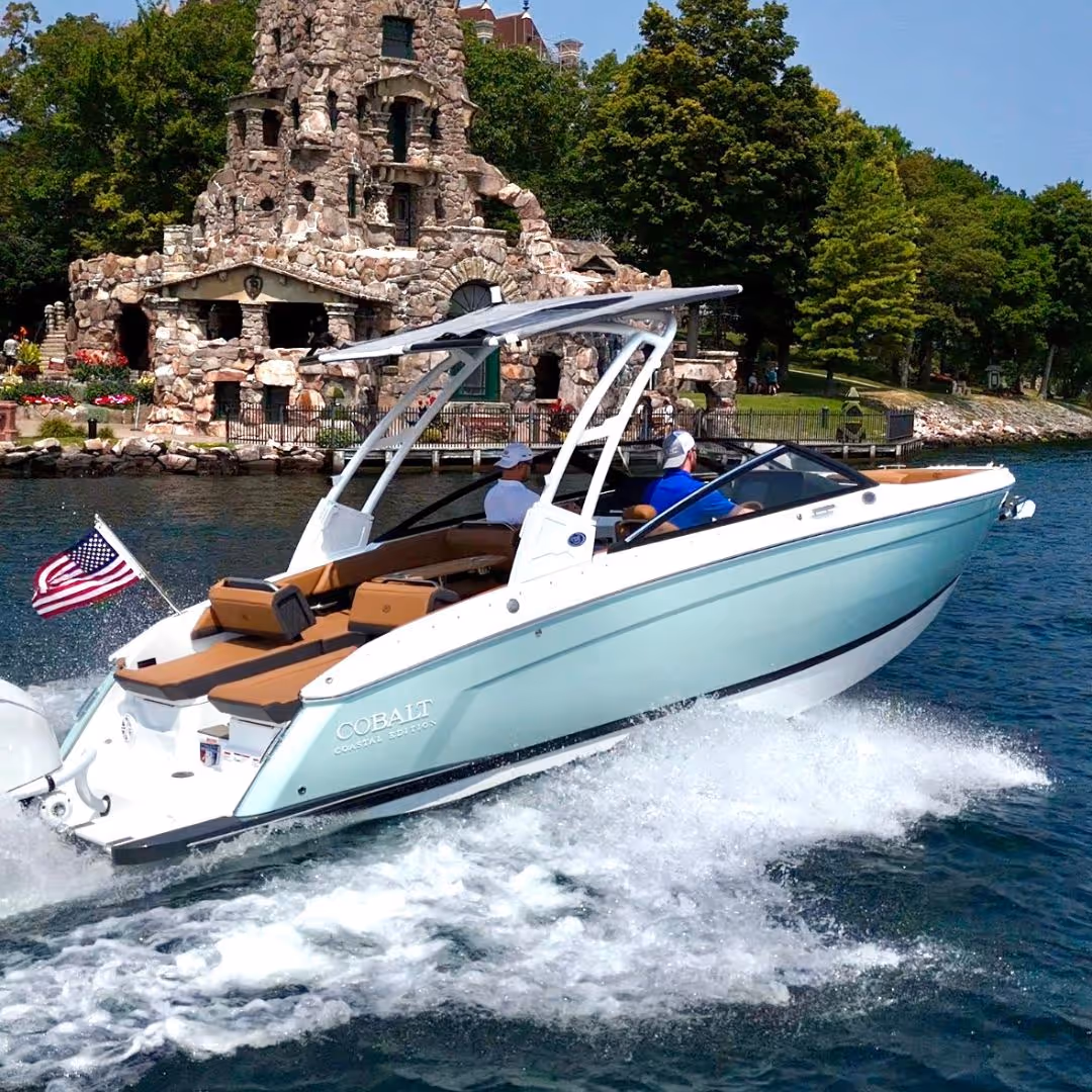 Light blue Cobalt Coastal Edition speedboat with two people cruising on water near a stone building and green trees.
