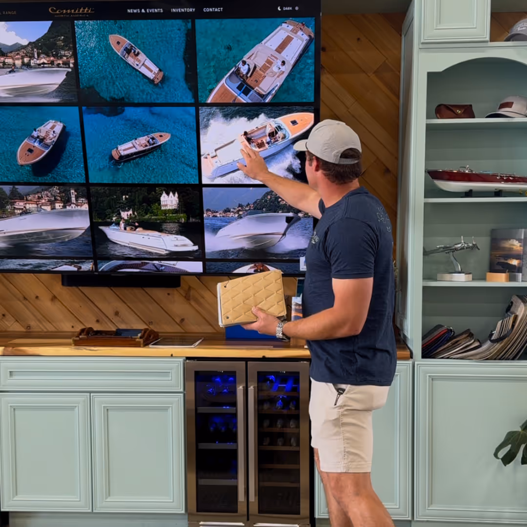 Man in casual clothing and cap pointing at a large screen displaying various luxury boats on water in a showroom or office setting.