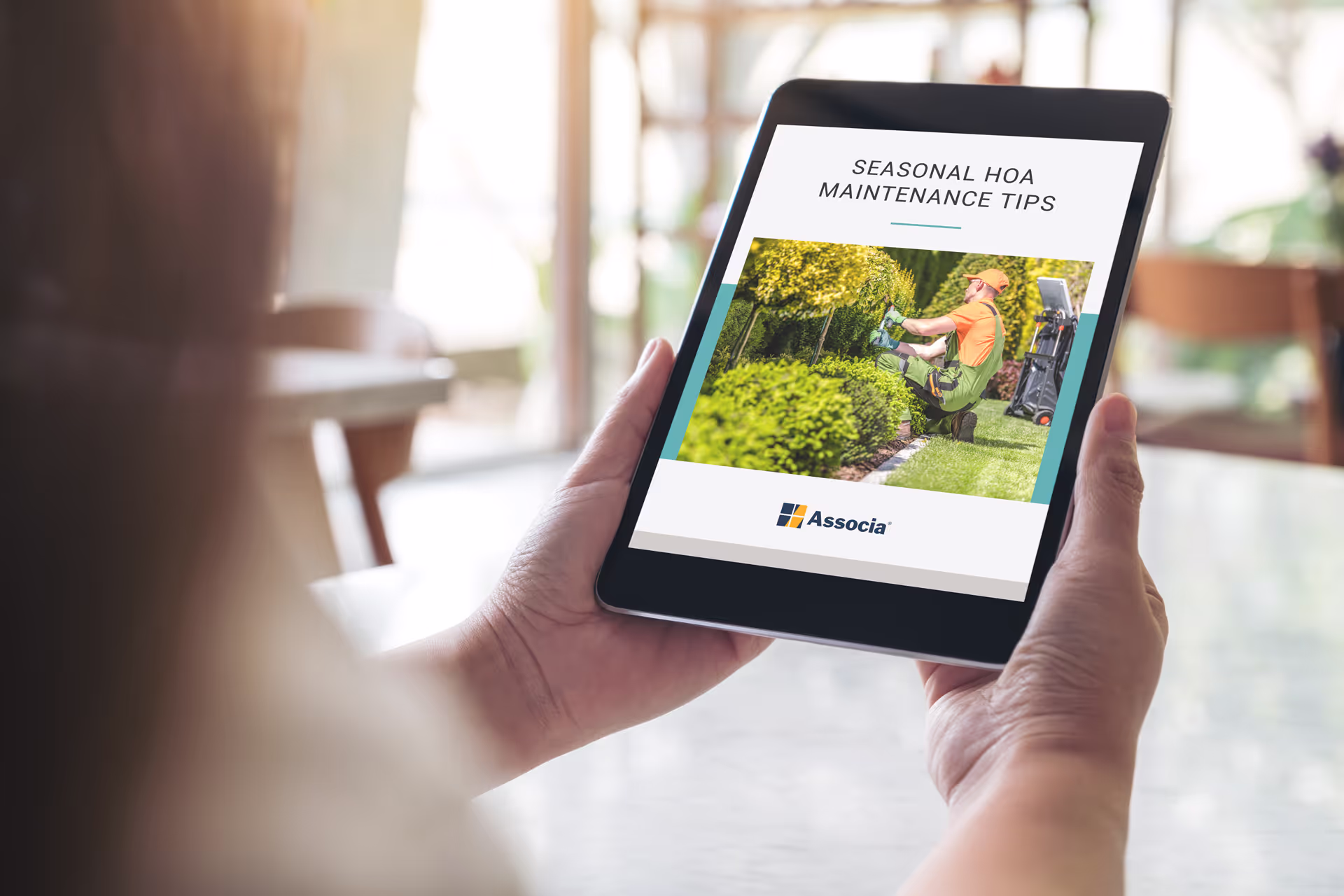 Associa eBook on tablet showing Seasonal HOA Maintenance Tips with landscaper trimming bushes in a residential garden