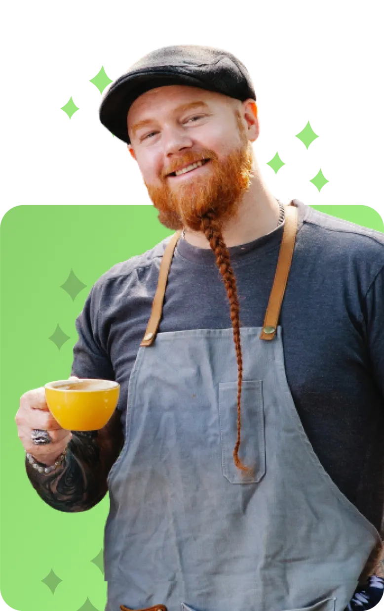 Smiling man with a reddish-brown beard, wearing a gray apron, dark shirt, black cap, holding a yellow coffee cup.