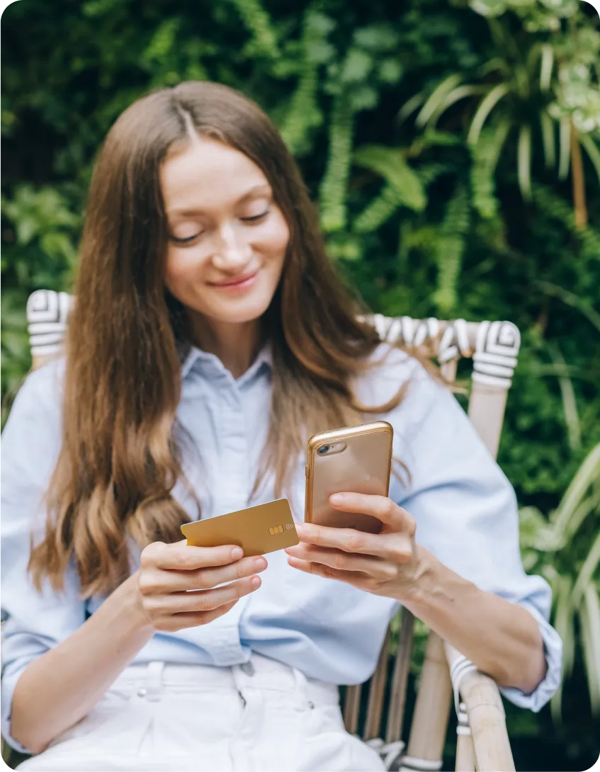 A woman is sitting outside, looking at her phone with a smile.