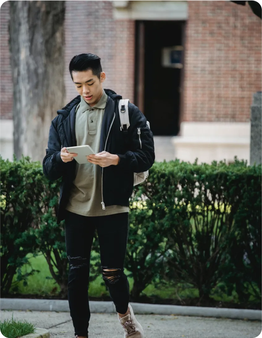 A person wearing a black jacket, green shirt, and carrying a backpack is walking while looking at a tablet. They are outdoors, with a brick building and bushes in the background.