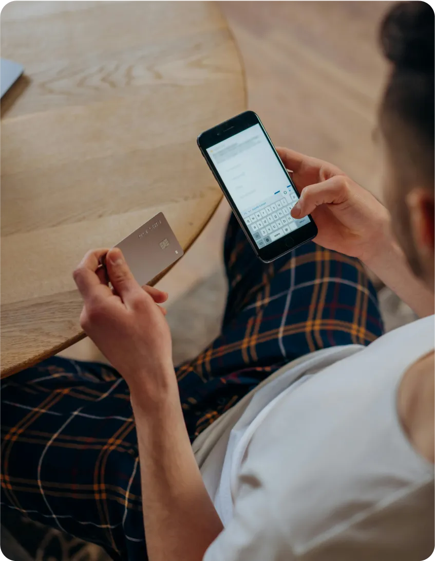 A person in a white shirt and plaid pants is seated at a round wooden table, holding a credit card in one hand and typing on a smartphone with the other, possibly engaged in online shopping or banking.