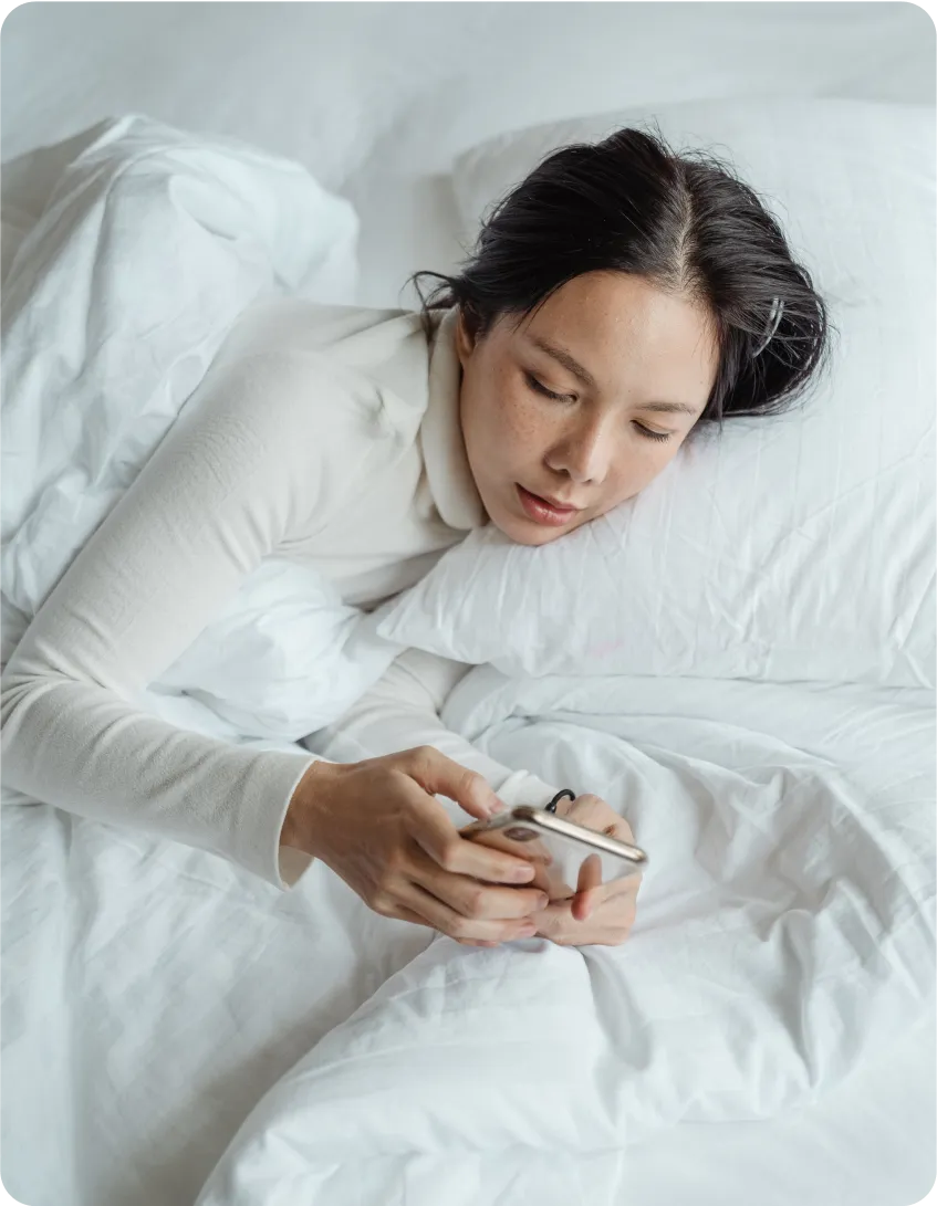 A person lying in bed under a white duvet, holding a smartphone. Their head rests on a pillow, and they appear to be looking at the screen. The bedding is crisp and white, matching the calm, relaxed atmosphere.