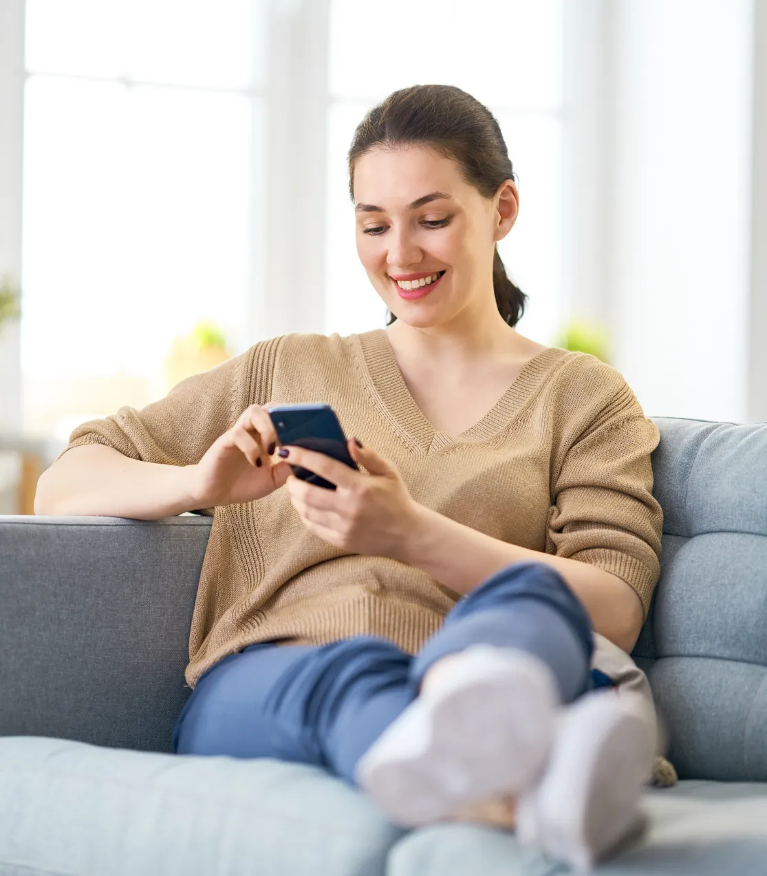 Woman sitting on a couch smiling while using a smartphone in a bright, cozy living room.