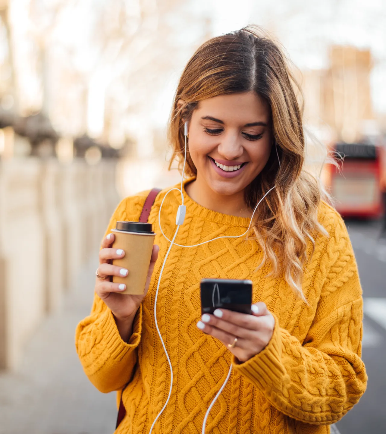 Woman in a yellow sweater smiling while listening to music on earphones and holding a coffee cup.