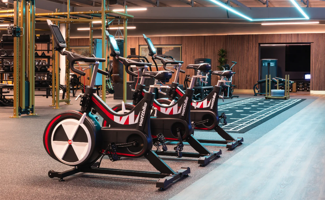 Indoor cycling area at David Lloyd St Neots, with decorated timber-clad wall and sprayed services overhead