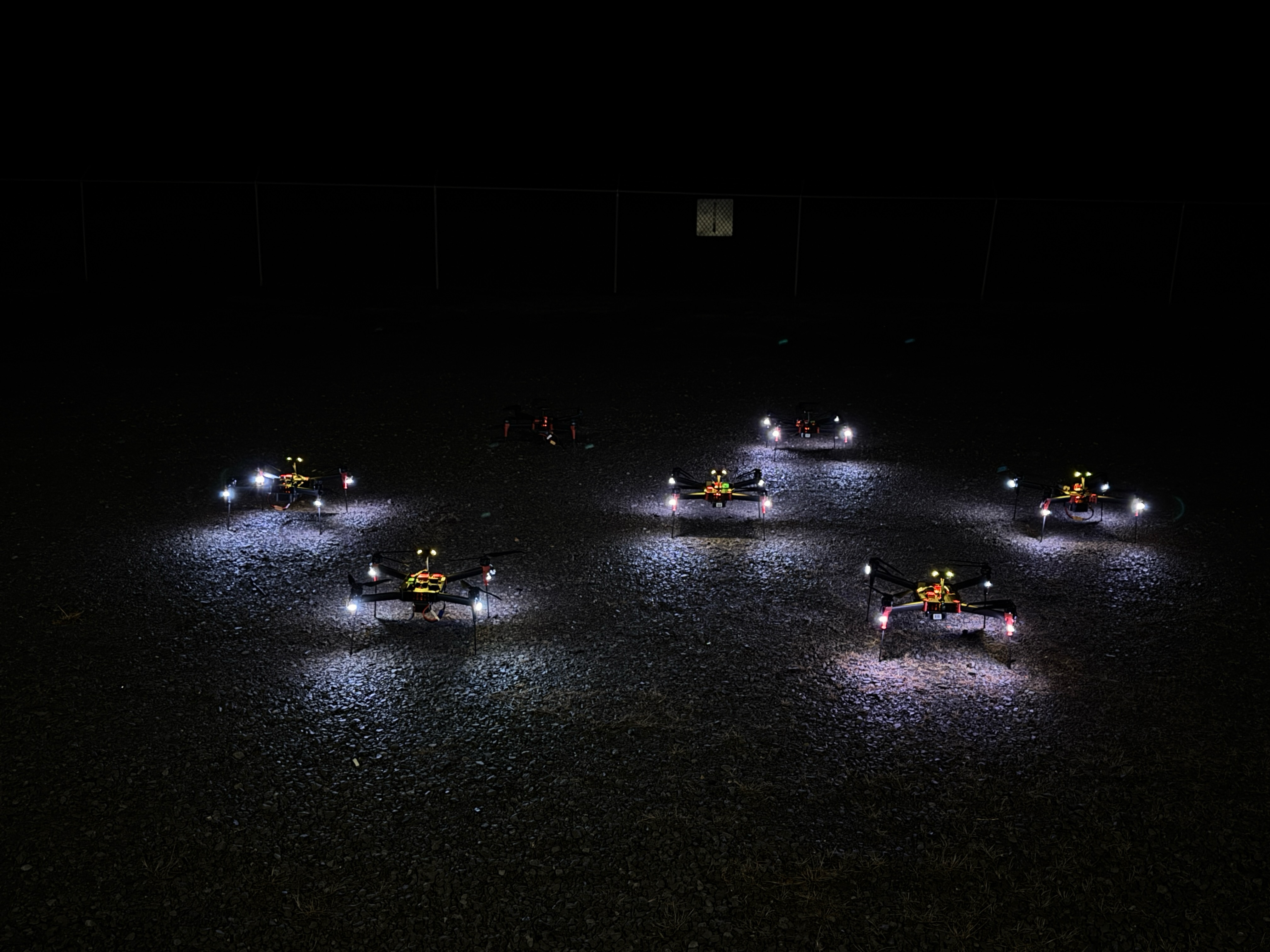 Seven drones with illuminated lights on a dark gravel surface at night.