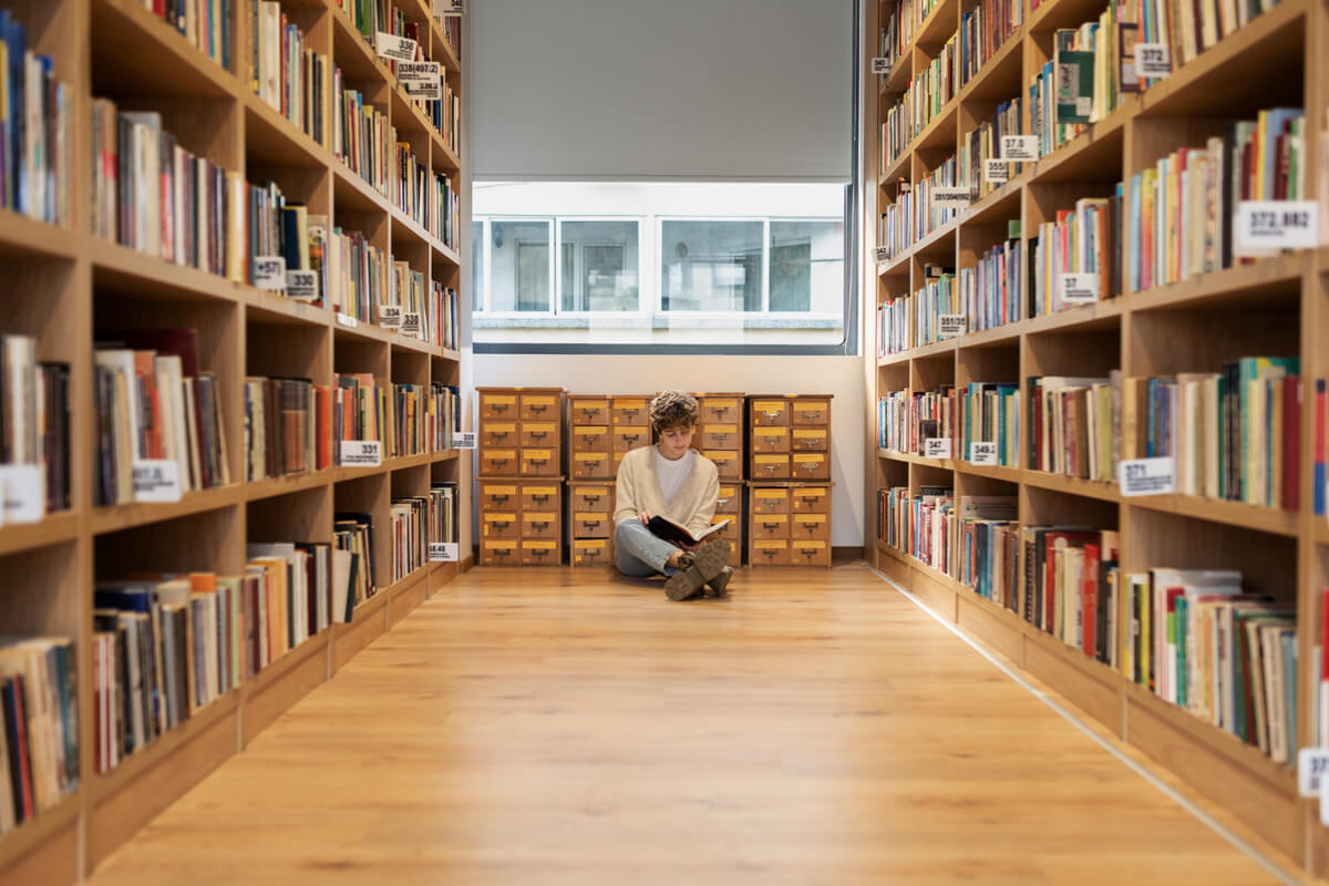 En person sitter på gulvet mellom bokhyller i et bibliotek og leser en bok