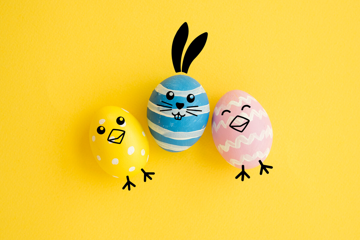 Tre påskegg dekorert som figurer, med gule og rosa egg som kyllinger og et blått egg som en påskehare mot gul bakgrunn