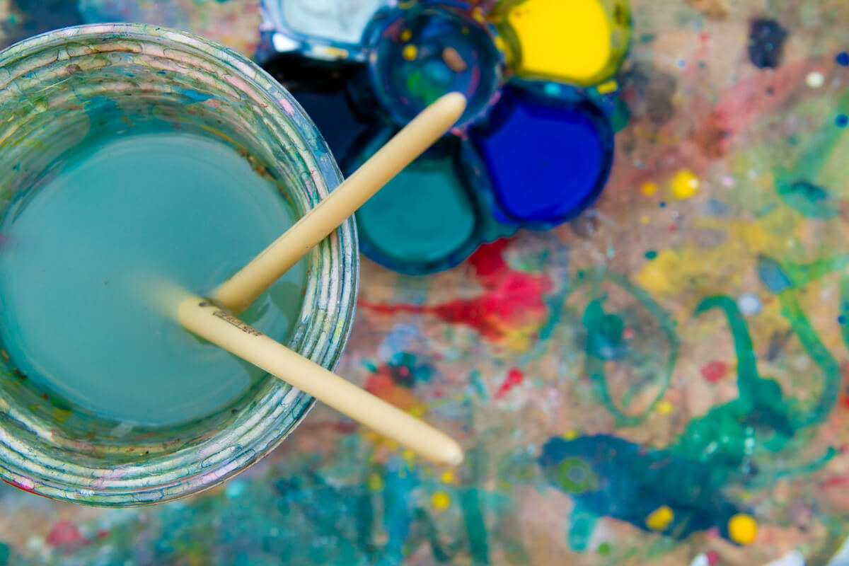 To malerpensler står i et glass med vann ved siden av en palett med sterke farger