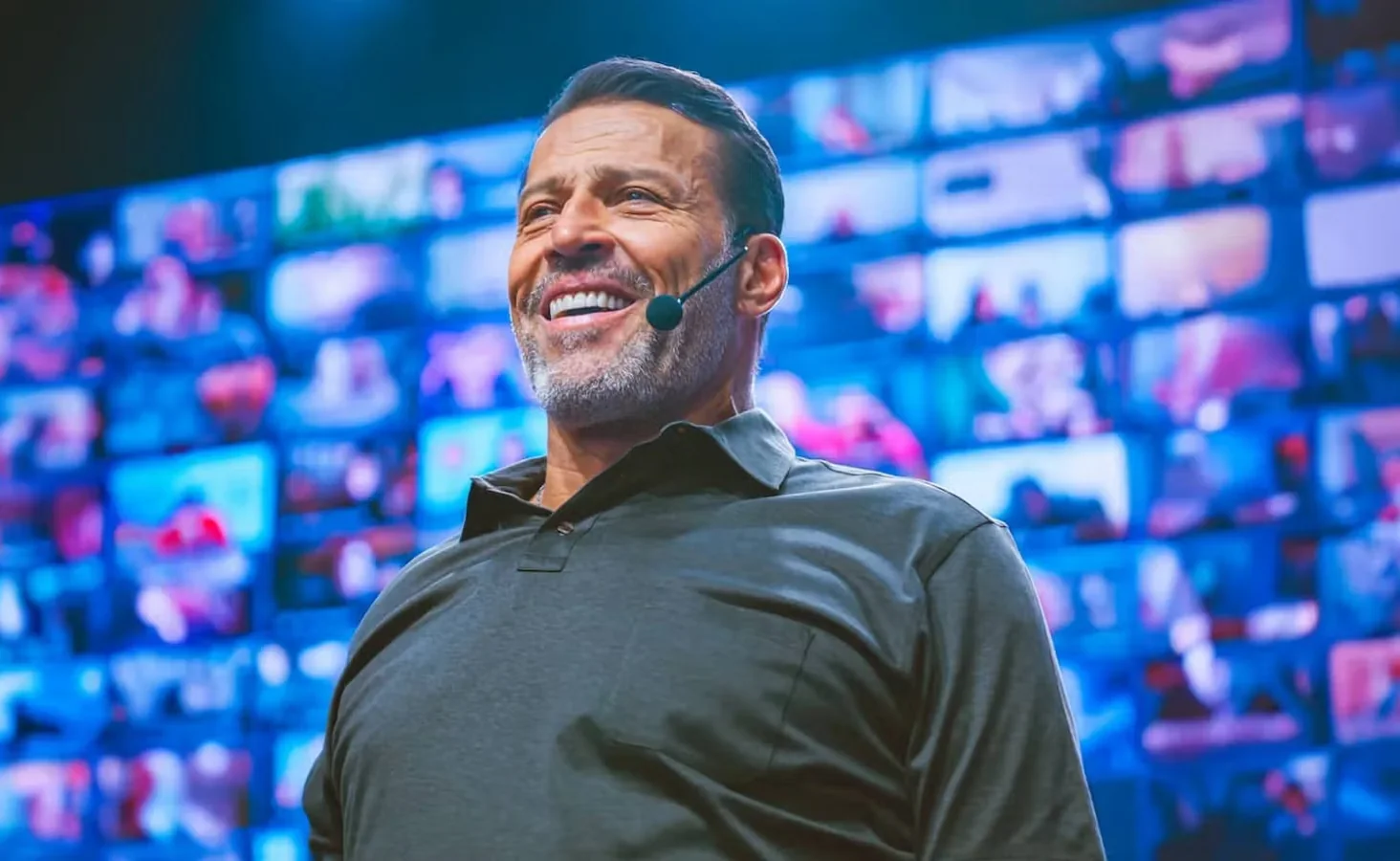 Smiling man wearing a headset microphone speaking in front of a large screen filled with small video call windows.