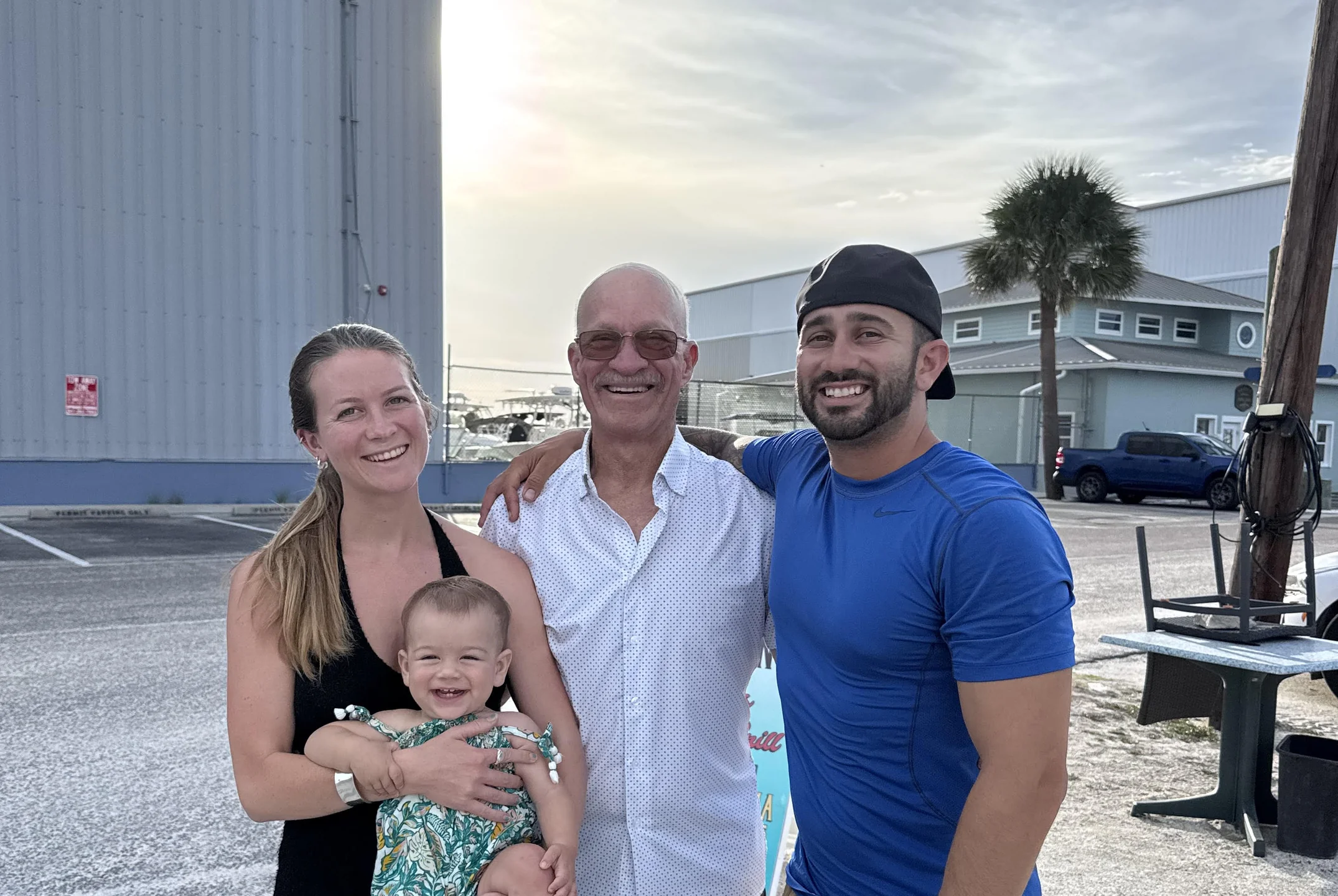 Smiling family of three adults and one baby standing outdoors near a marina with boats and buildings in the background.