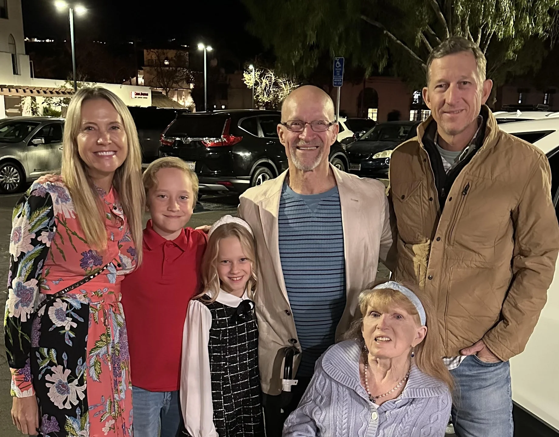 Group of six people smiling outdoors at night in a parking lot, including two children and four adults, one woman seated in a wheelchair.