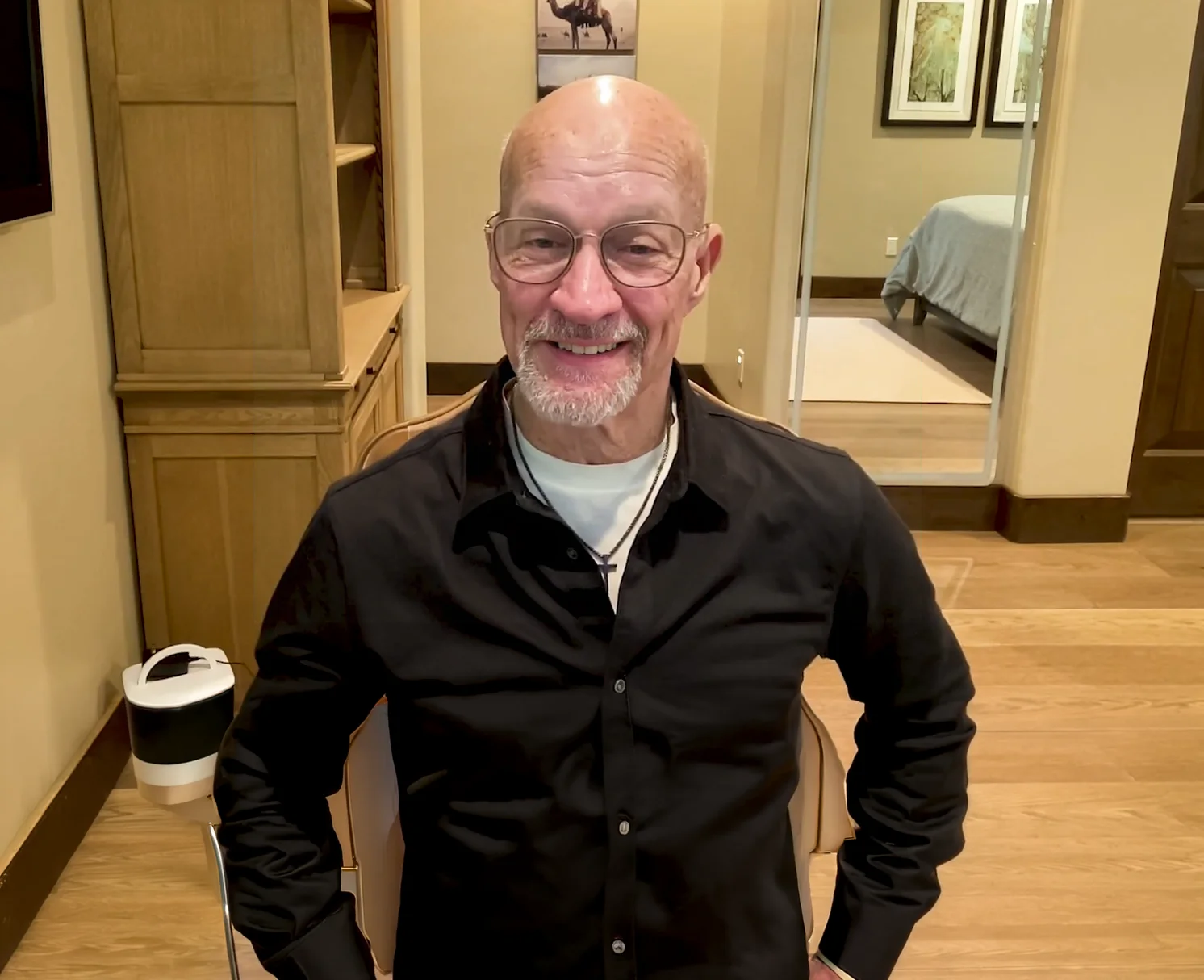 Smiling bald man with glasses and a goatee wearing a black shirt sitting indoors with wooden furniture and a bed visible in the background.