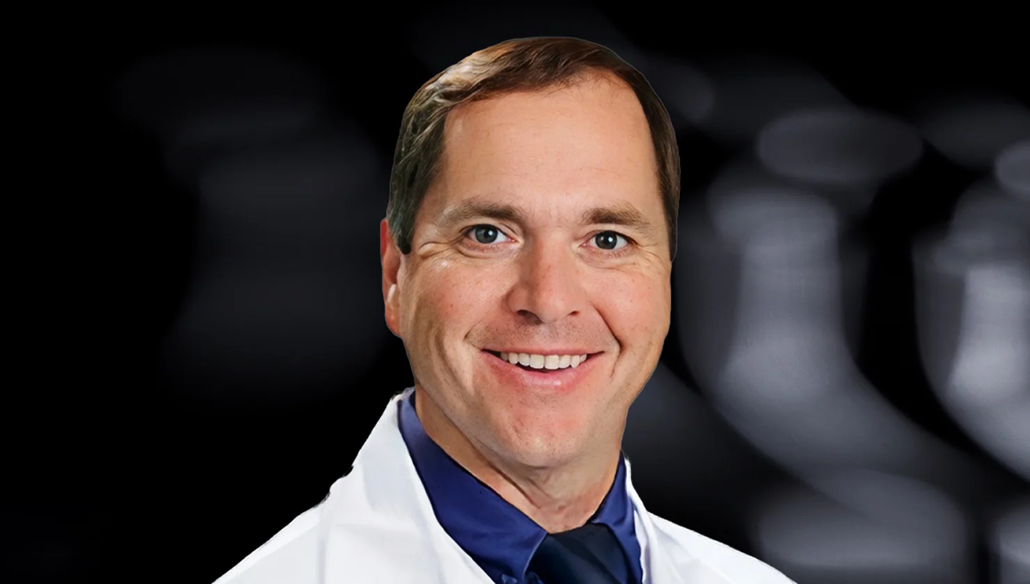 Smiling man wearing a white lab coat with a blue shirt and tie against a dark blurred background.