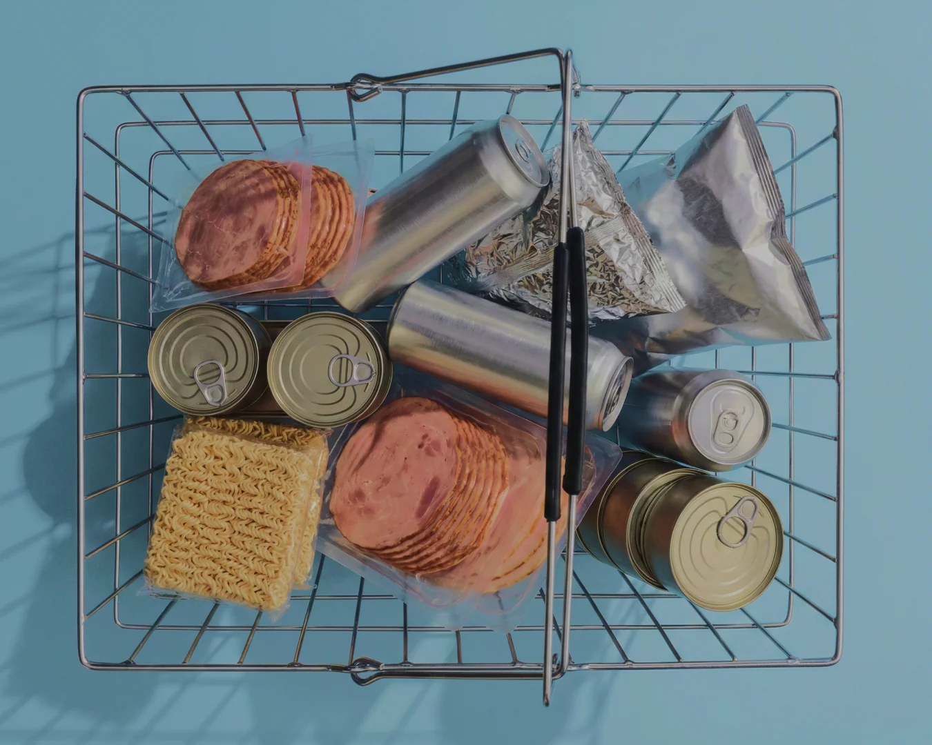 Shopping basket with canned goods, packs of sliced processed meat, instant noodle blocks, aluminum beverage cans, and silver foil snack bags on a blue background.