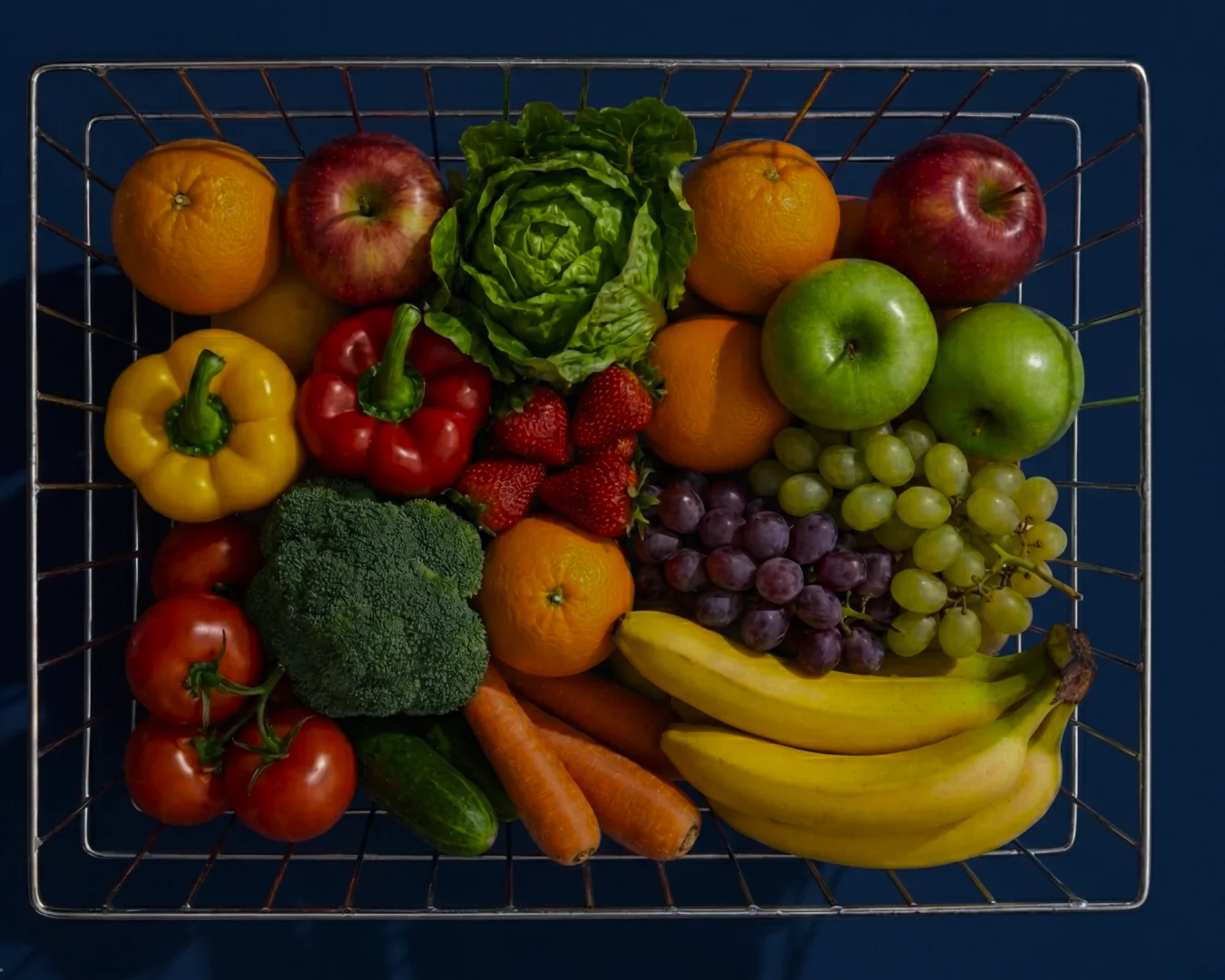 Metal basket filled with assorted fresh fruits and vegetables including bananas, grapes, oranges, apples, strawberries, carrots, broccoli, tomatoes, bell peppers, cucumber, and lettuce.