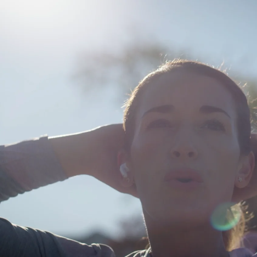 Close-up of a woman outdoors with hands behind her head, wearing wireless earbuds, backlit by the sun.