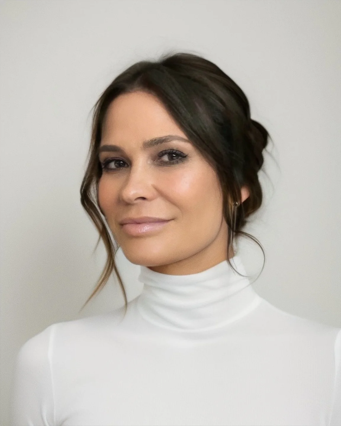 Woman with dark brown hair styled in a loose updo, wearing a white turtleneck and subtle makeup, smiling slightly against a plain gray background.