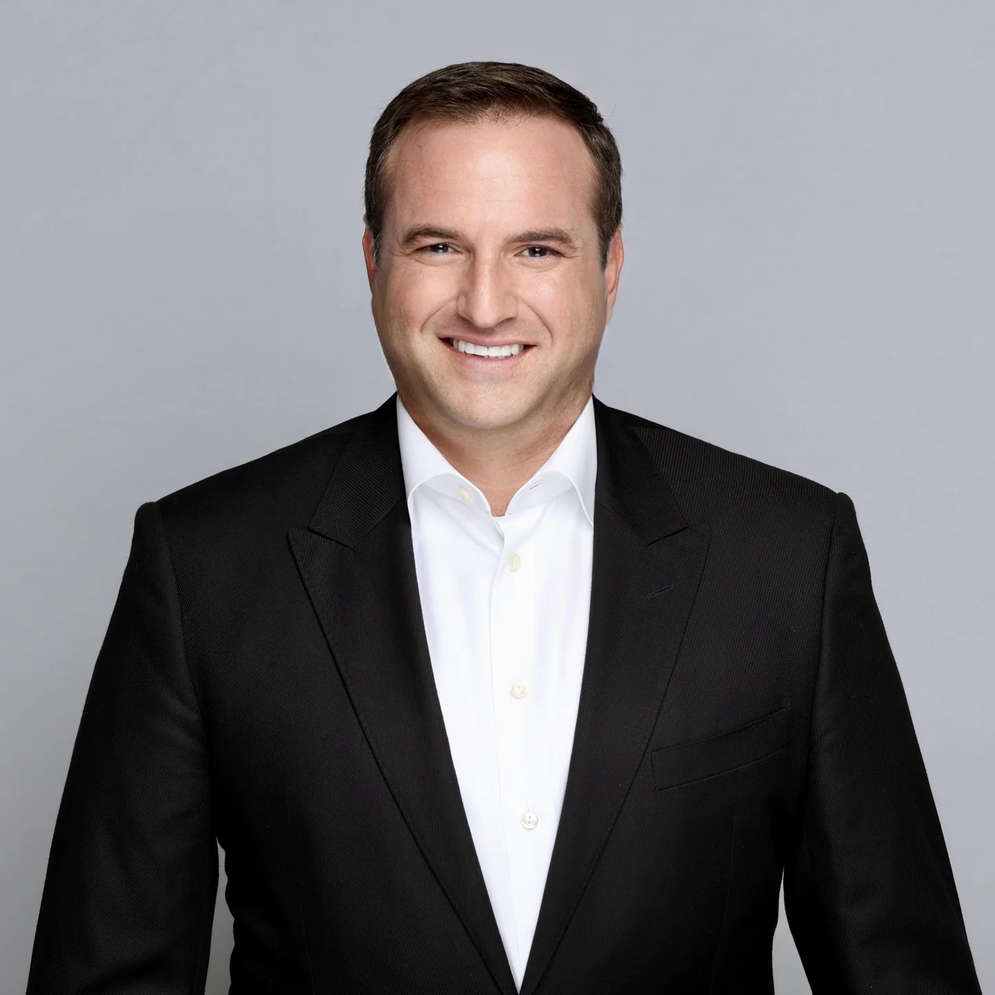 Smiling man with short brown hair wearing a black suit jacket and white shirt against a plain gray background.