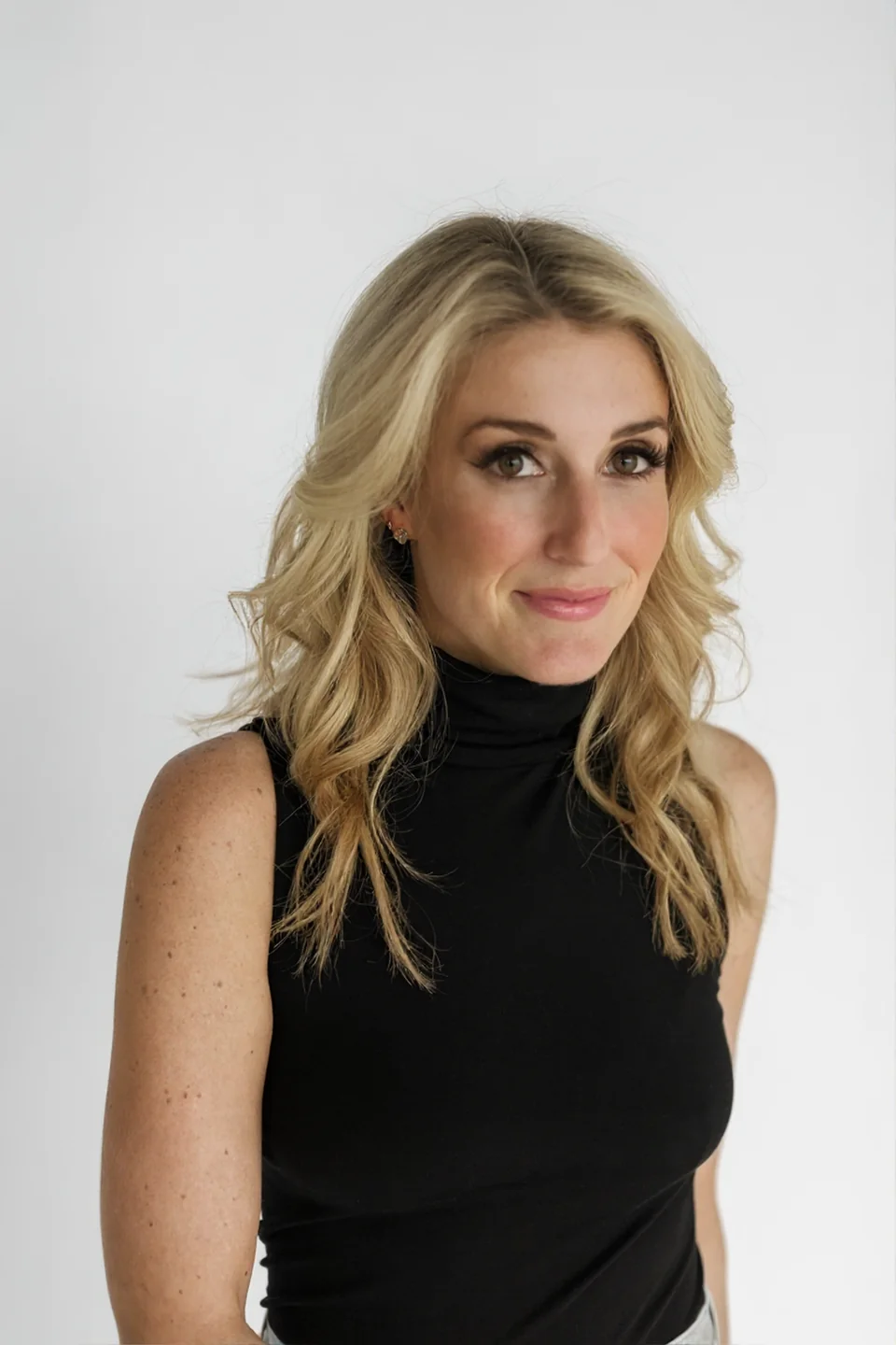 Blonde woman with wavy hair wearing a black sleeveless turtleneck against a plain light background.