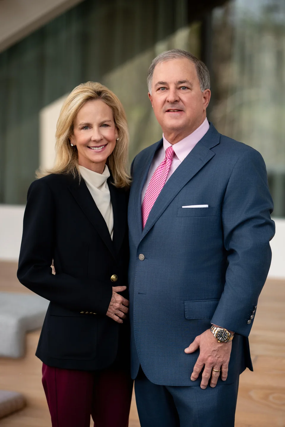 Smiling couple dressed formally, the woman in a black blazer and burgundy pants, and the man in a blue suit with a pink tie.
