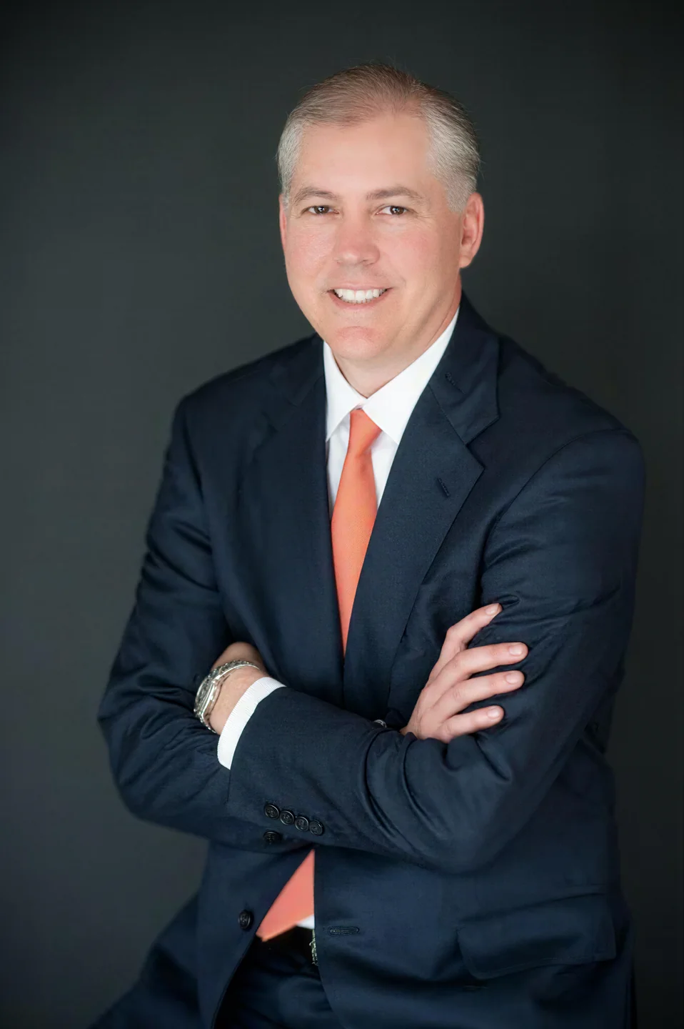 Smiling middle-aged man in a dark suit with an orange tie, arms crossed against a dark background.