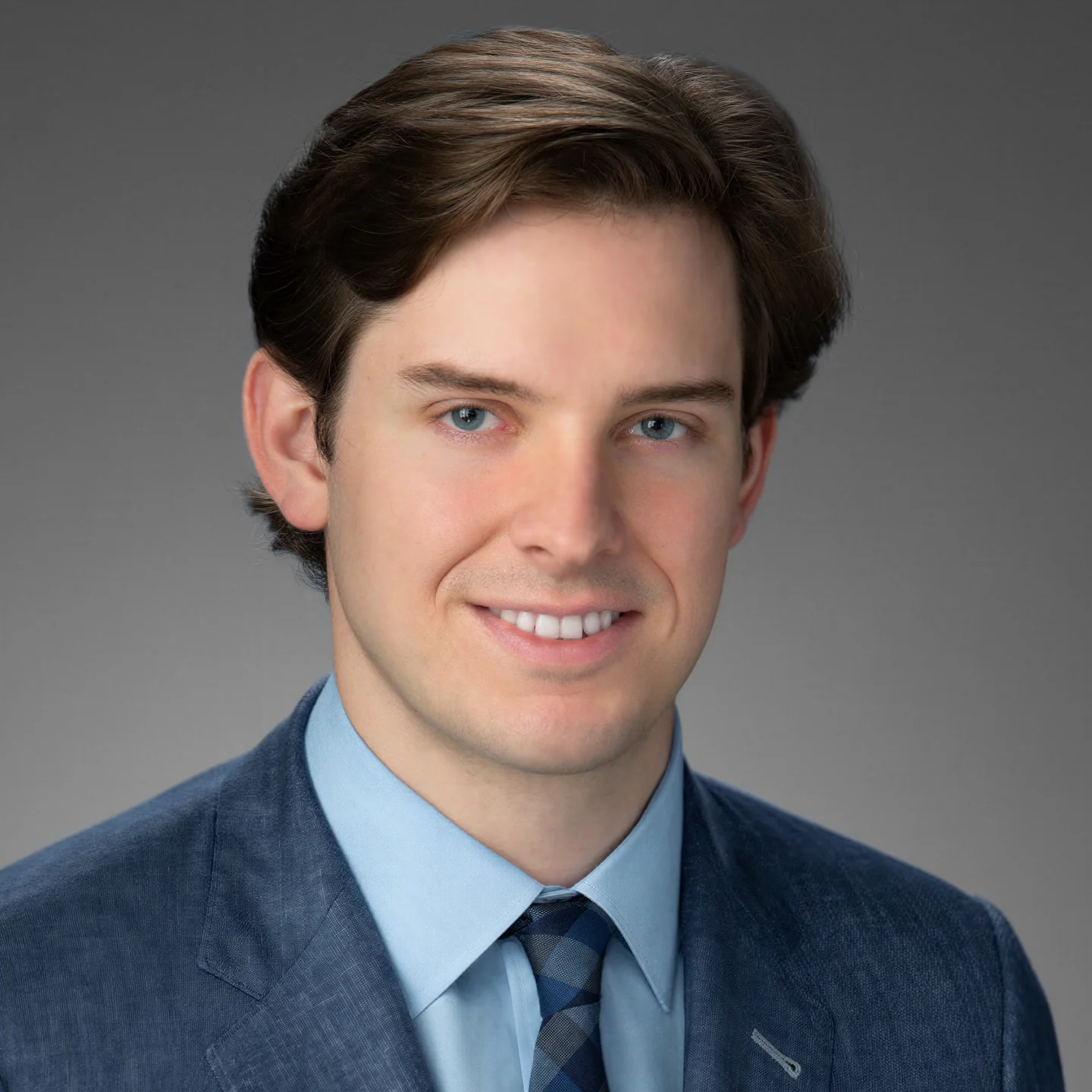 Smiling young man with brown hair wearing a blue suit, light blue shirt, and a blue plaid tie against a gray background.