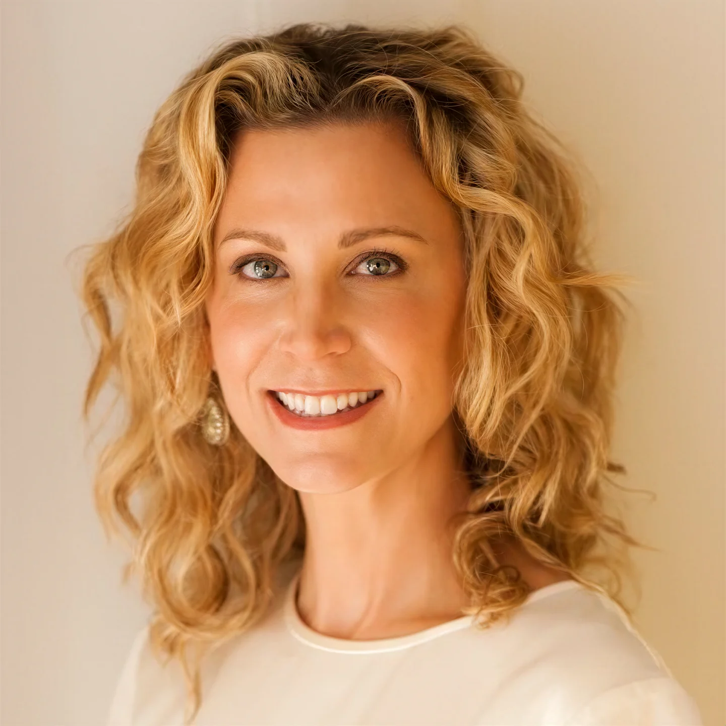 Smiling woman with curly blonde hair wearing a white top and dangling earrings against a beige background.