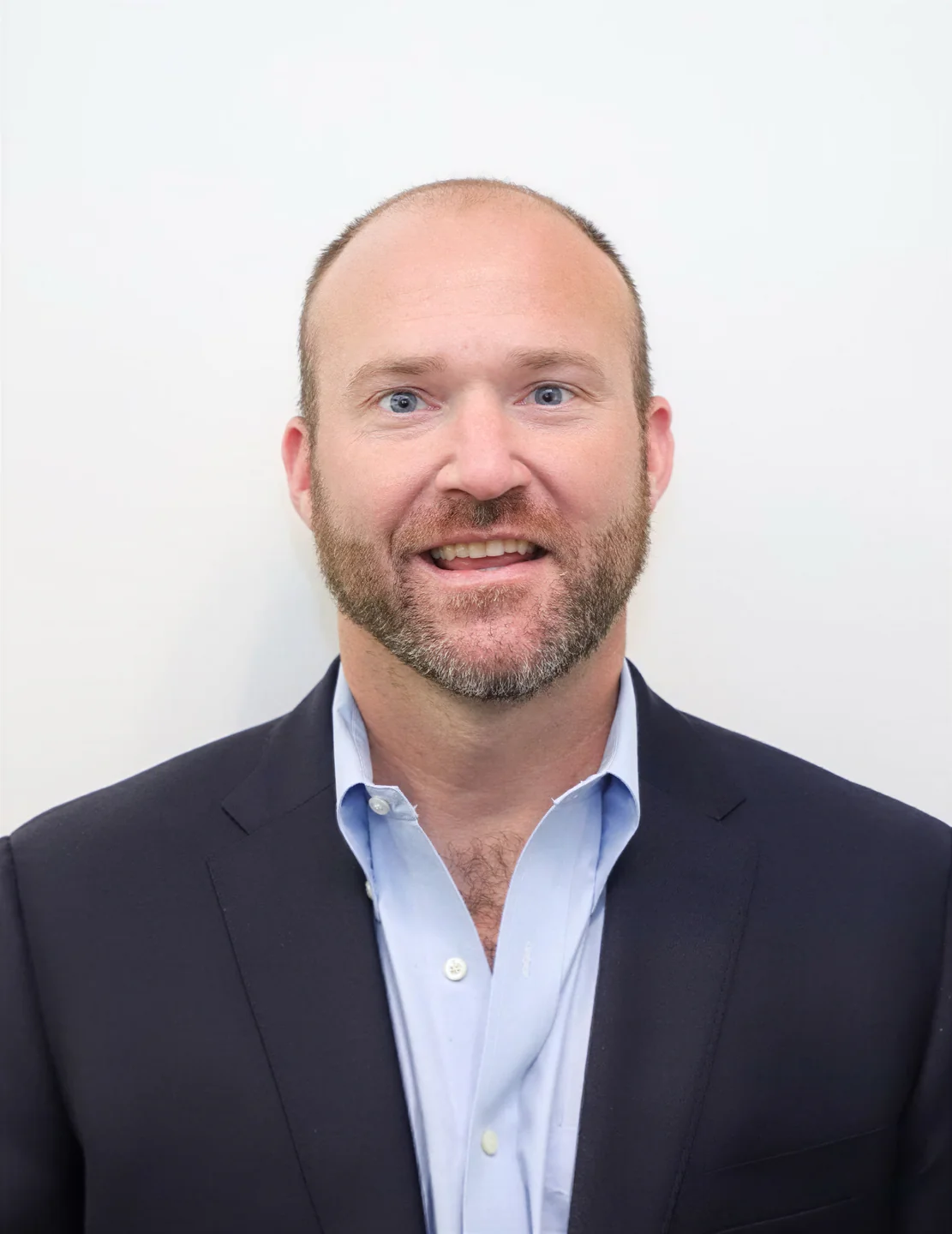 Smiling bald man with a short beard wearing a blue dress shirt and dark blazer against a plain white background.