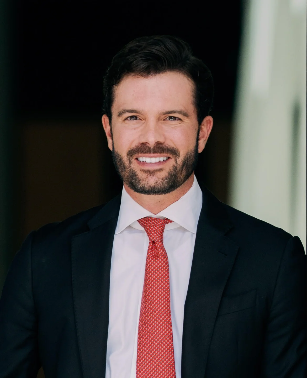 Smiling man with dark hair and beard wearing a black suit jacket, white shirt, and red patterned tie.