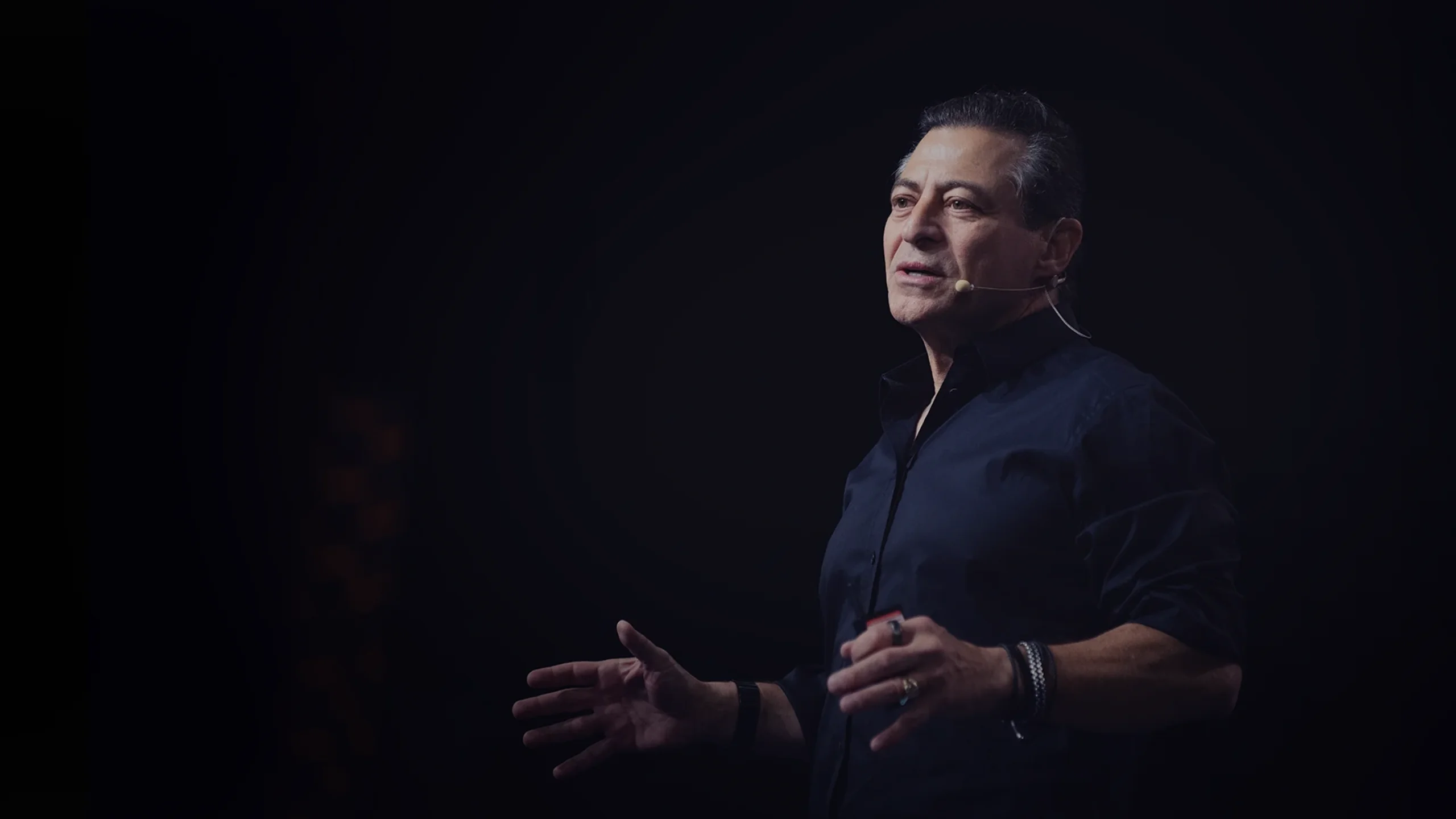Man wearing a dark shirt speaking with a microphone headset against a black background.