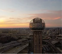 Dawn view of the Reunion Tower with its illuminated geodesic dome atop a tall cylindrical base, overlooking highways and city buildings under a soft orange and blue sky.